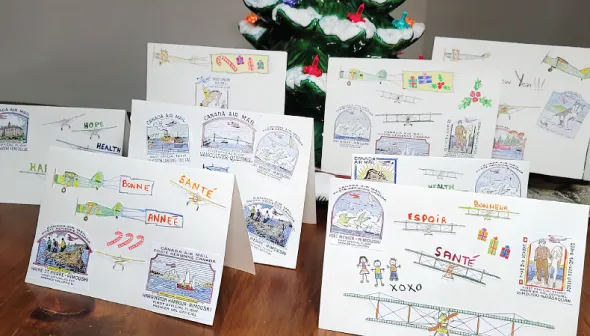 Eight brightly-coloured holiday cards are arranged on a wooden tabletop; each card features different aviation-themed images. Christmas décor is visible in the foreground and background of the image.