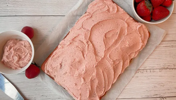 Une vue aérienne montre un gâteau rectangulaire avec du glaçage rose, reposant sur un plateau placé sur une surface de bois. Un petit bol de glaçage, un petit bol de fraises et une spatule à glaçage sont visibles près du plateau.
