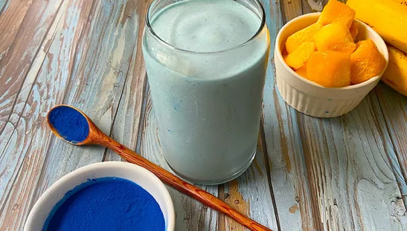 Un verre rempli d’une épaisse boisson bleu repose sur une surface de bois rustique. Un bol et une cuillère avec de la poudre bleue se trouvent à l'avant-plan, avec des mangues et des bananes à l'arrière-plan.