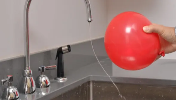 A balloon is held near a sink with running water. The stream of the water bends toward the balloon.