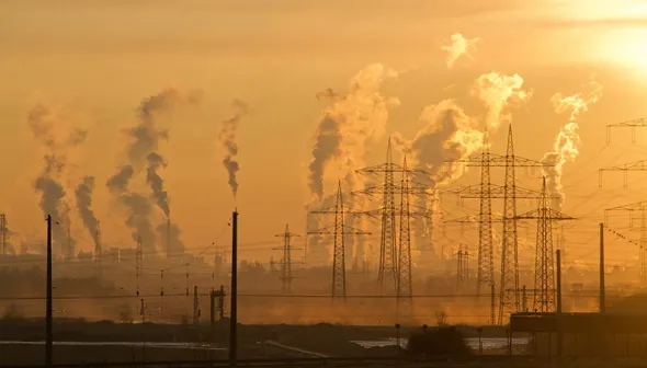 Des poteaux téléphoniques et des pylônes électriques sont au premier plan et une ligne d'horizon brumeuse avec de multiples cheminées rejetant de la pollution dans un ciel doré à l'arrière-plan.
