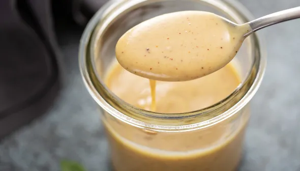 Une cuillère remplie de sauce moutarde et miel repose sur le dessus d’un petit pot ouvert.