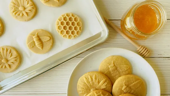 Des biscuits sont disposés sur une plaque à biscuits, et d’autres dans une assiette blanche posée sur une surface blanche. À côté, on voit un pot de miel et une cuillère à miel.