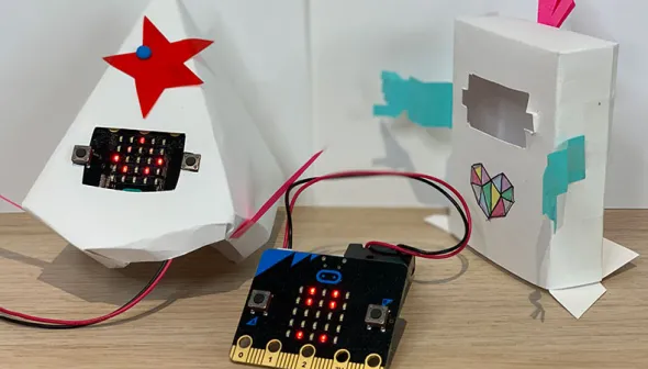 A square micro:bit device place between, on the left a pyramid-shaped paper sculpture adorned with a red star in which a micro:bit is displaying a happy smile, and on the right a rectangle sculpture decorated with a colourful heart with an empty space for a micro:bit display. 