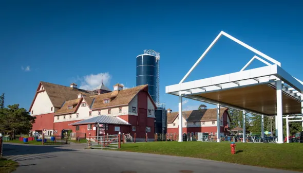 A wide shot of two large, red-and-white agricultural buildings and a navy blue silo. Green grass and a covered, outdoor picnic area are visible in the foreground, and a blue sky in the background.