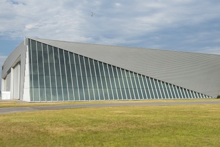 Photo du hangar de réserve du musée, un grand bâtiment gris avec un grand triangle de fenêtres sur la façade.  Le bâtiment se détache sur un ciel bleu parsemé de nuages et un champ d'herbe verte.