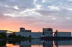 A ship in front of a large grain elevator; the sun rises in the background.
