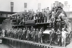 Photographie en noir et blanc d’employés qui se tiennent debout sur une locomotive à vapeur et devant celle-ci. On dénombre une quarantaine d’hommes.