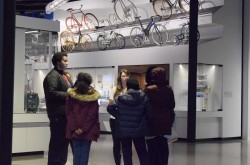 A group of teenagers and one coordinator gather in a circle to sort out their ideas and problem solve. Behind them is a showcase of the different kinds of bikes there have been through time. 