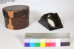 Un petit prisme en métal et en verre repose à côté de sa vieille boîte de rangement brune sur un fond blanc. Une charte de couleur Kodak se trouve devant les artefacts.