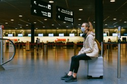 A young woman sits on her luggage in an empty airport terminal, gazing off into the distance to the left of screen. She is alone and wearing a blue medical mask, with headphones draped around her neck.
