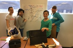 Four students, two in green shirts, two in grey shirts, stand on both sides of a poster with their invention drawn on it. Materials for building their invention are scattered in front of them. 