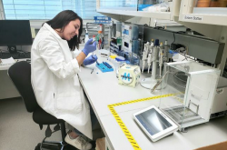 A young woman wearing a white lab coat and blue rubber gloves sits in a laboratory, working with a variety of tools in front of her. 