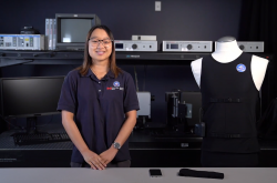 A young woman wearing a navy blue shirt smiles as she stands next to a mannequin dressed in a black tank top. A computer and a variety of equipment is visible on shelves in the background.