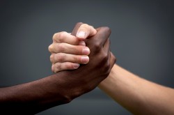 Two hands clasped together, one Black and one white, against a dark grey background. 