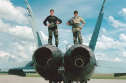 Two servicewomen in flight suits pose confidently atop a fighter jet at an airstrip on a clear blue day.