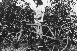 A black-and-white photo of a man standing outside with his left hand on the seat of a wooden bicycle.