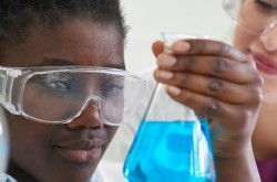 A close-up shot of two young people wearing safety goggles, in a lab environment. The person in the foreground is holding up a glass beaker filled will blue liquid.