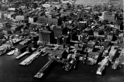 A black and white photograph taken from the air. The photo shows a series of docks with buildings on the port, and city streets behind. The Halifax Citadel is in the background.