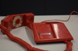 A red plastic telephone with the handset off of the base on a light grey table. There are scratches on the phone which is an angular design. The rotary dial is on the handset and attached to the base by a red spiral cord. 