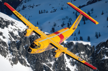 Avion Buffalo CC-115 jaune et rouge en vol, sur fond de montagnes enneigées.